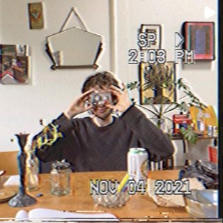 Jasper Craig Adams holds a vintage camera to his eye, obscuring his face. He is wearing a grey jumper, sitting at a wooded dining table where an assortment of objects are placed - a candleholder, a jar of plant cuttings, a can of drink, and some papers. Behind him a white wall features various artworks and a hanging mirror. The image is captured as if from a digital camera still, complete with slight pixelation and stamped with the time and date with white digital text