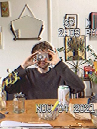 Jasper Craig Adams holds a vintage camera to his eye, obscuring his face. He is wearing a grey jumper, sitting at a wooded dining table where an assortment of objects are placed - a candleholder, a jar of plant cuttings, a can of drink, and some papers. Behind him a white wall features various artworks and a hanging mirror. The image is captured as if from a digital camera still, complete with slight pixelation and stamped with the time and date with white digital text