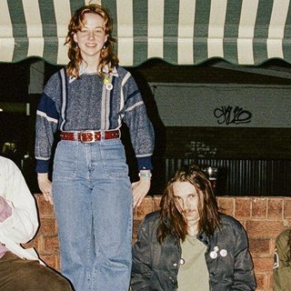 A film photograph of the four band members of R.M.F.C. They are sitting (and one is standing) against a brickwall, with a green and white striped awning above them.