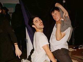 Marcus Whale is on the left, bent to his knees and arching back with a gleeful look his face He is wearing a white t shirt and grey striped pants. Gus McGrath is to his right, legs spread wide and his arms in a cross over his head. He wears a white shirt, black pants, and a grey jumper as a shawl over his head. His expression is of a slight smile. They are at an event where there is a crowd and live music.