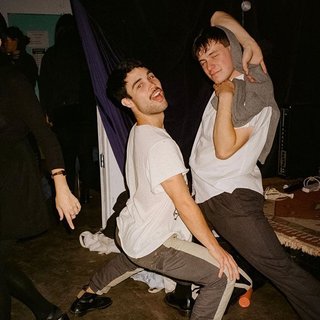 Marcus Whale is on the left, bent to his knees and arching back with a gleeful look his face He is wearing a white t shirt and grey striped pants. Gus McGrath is to his right, legs spread wide and his arms in a cross over his head. He wears a white shirt, black pants, and a grey jumper as a shawl over his head. His expression is of a slight smile. They are at an event where there is a crowd and live music.