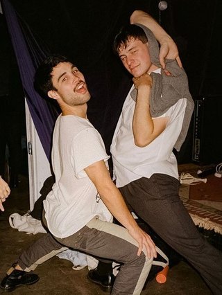 Marcus Whale is on the left, bent to his knees and arching back with a gleeful look his face He is wearing a white t shirt and grey striped pants. Gus McGrath is to his right, legs spread wide and his arms in a cross over his head. He wears a white shirt, black pants, and a grey jumper as a shawl over his head. His expression is of a slight smile. They are at an event where there is a crowd and live music.