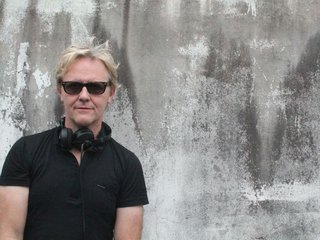 Stephen looks directly at the camera, wearing a black tee, black sunglasses and headphones around his neck. He is backgrounded by a concrete wall.