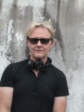 Stephen looks directly at the camera, wearing a black tee, black sunglasses and headphones around his neck. He is backgrounded by a concrete wall.