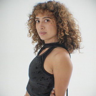 Tangela looks past her left shoulder into the camera with an inquisitive look. She is wearing a strappy black top. She stands in front of a plain white background and their is a pixelated filter over the image .