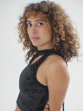 Tangela looks past her left shoulder into the camera with an inquisitive look. She is wearing a strappy black top. She stands in front of a plain white background and their is a pixelated filter over the image .
