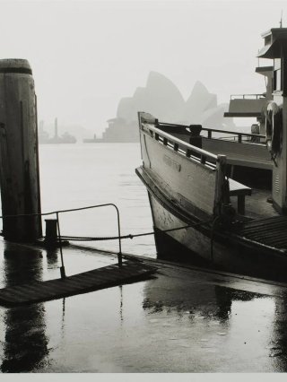 A black and white photo of a ferry docked on the water, with a hazy sydney opera house in the background