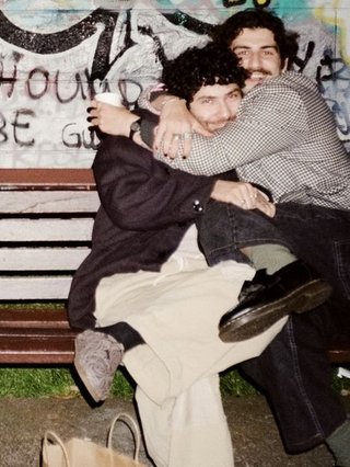 Luke is sitting with his legs crossed in the tight embrace of his friend Chris, on a bench in Camperdown Park. They are both smiling at the camera and wearing warm jackets and pants. Behind them a graffiti'd wall shows large graphics and cut off text.