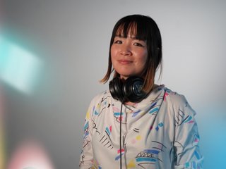 Kana Frazer is smiling at the camera with her head tilted slightly to her left. She wears big black headphones around her neck, and a white hoodie with colourful illustrations on it. She stands in front of a grey background, and colourful light beams border the left.