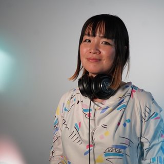Kana Frazer is smiling at the camera with her head tilted slightly to her left. She wears big black headphones around her neck, and a white hoodie with colourful illustrations on it. She stands in front of a grey background, and colourful light beams border the left.