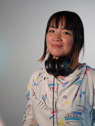Kana Frazer is smiling at the camera with her head tilted slightly to her left. She wears big black headphones around her neck, and a white hoodie with colourful illustrations on it. She stands in front of a grey background, and colourful light beams border the left.