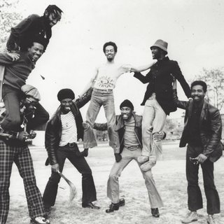 The image is a black and white photo of the 8 members of the band Cymande standing outside. Some members are standing on each others shoulders and legs in a human tower.