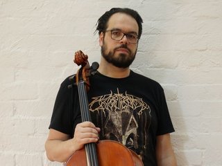 Peter holds a cello, wearing a black graphic tee and glasses. He looks directly at the camera against a white bricked wall.