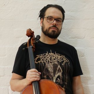Peter holds a cello, wearing a black graphic tee and glasses. He looks directly at the camera against a white bricked wall.