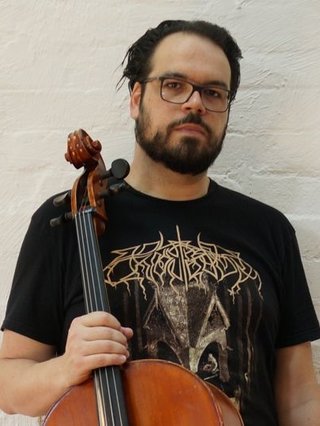 Peter holds a cello, wearing a black graphic tee and glasses. He looks directly at the camera against a white bricked wall.