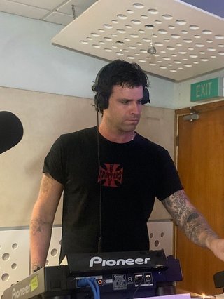 A tall man wearing a dark shirt behind a dj controller in a studio.