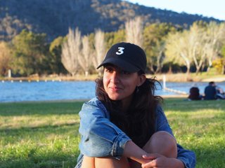 Kanika Kirpalani sits cross legged on the grass, her arms hugging her knees. She wears a denim jacket and a cap with the number 3 embroidered on it. She is in a grassy field with a river behind her and dense forest and mountain in the background.