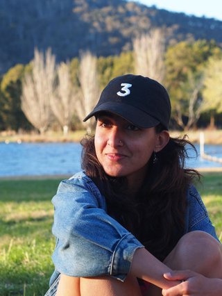 Kanika Kirpalani sits cross legged on the grass, her arms hugging her knees. She wears a denim jacket and a cap with the number 3 embroidered on it. She is in a grassy field with a river behind her and dense forest and mountain in the background.