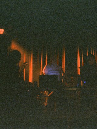 A grainy film image of shadowy band figures on stage, with red light behind them