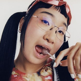 Yaeji looks to the camera. She is standing against a white backdrop with a lollipop in her mouth.